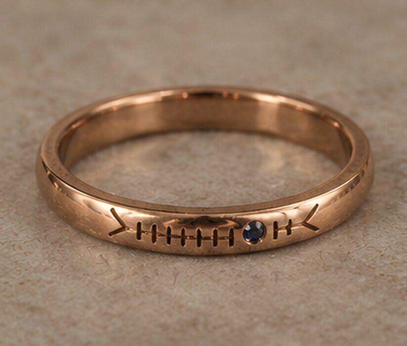 A medium rose gold band with a name cut in Ogham along the outside and a small sapphire set into the line