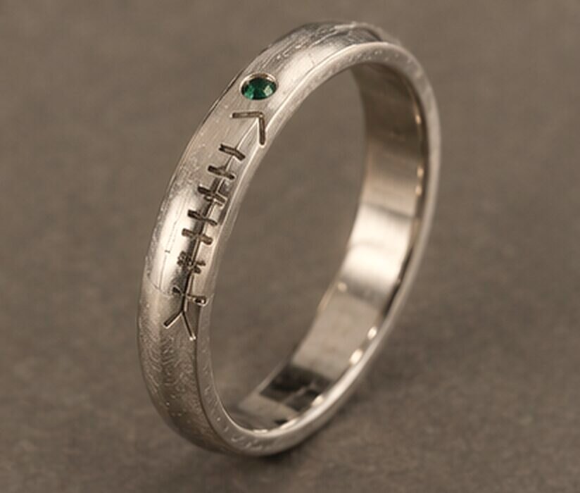 A thick white gold band with a name cut in Ogham along the outside and a small green tourmaline set into the line