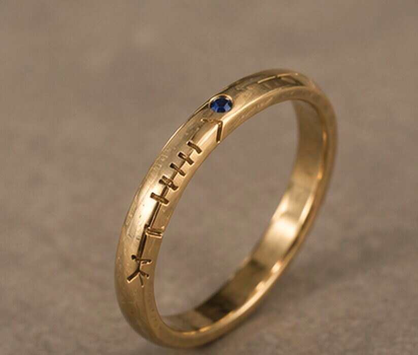 A thin yellow gold band with a name cut in Ogham along the outside and a small sapphire set into the line