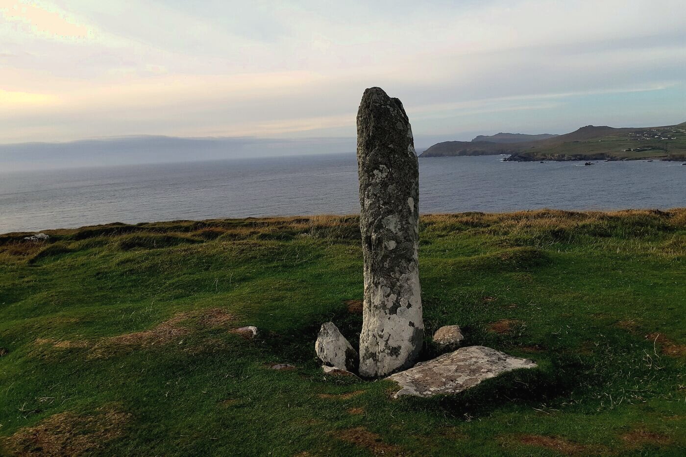 A tall weathered standing stone on a grassy headland, with the sea behind it