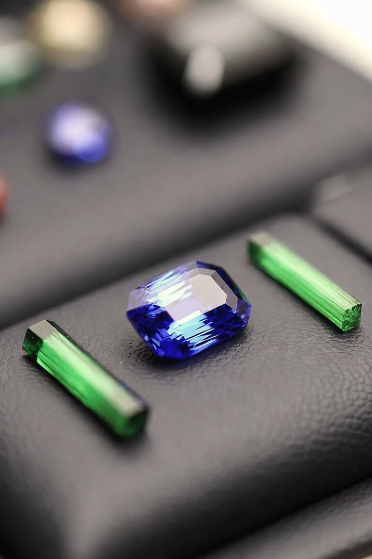 A cut blue sapphire between two long green tourmaline crystals on a dark leather tray